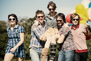 Grupo com óculos de sol em parque. Dois homens levantam uma mulher no centro da imagem.