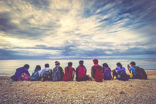 Grupo de pessoas com mochilas sentado lado a lado na areia a beira-mar.