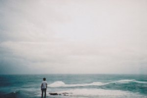 Homem em pé na beira do mar de costas olhando as ondas quebrarem
