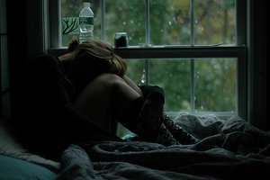 Pessoa sentada na cama com cabeça abaixada e garrafa d'água e remédios na janela