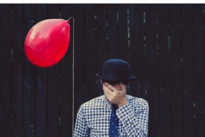 Homem segurando balão vermelho com mão no rosto e chapéu