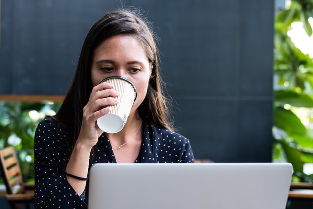 Mulher bebendo café em um copo de papel, enquanto olha para a tela de um notebook.