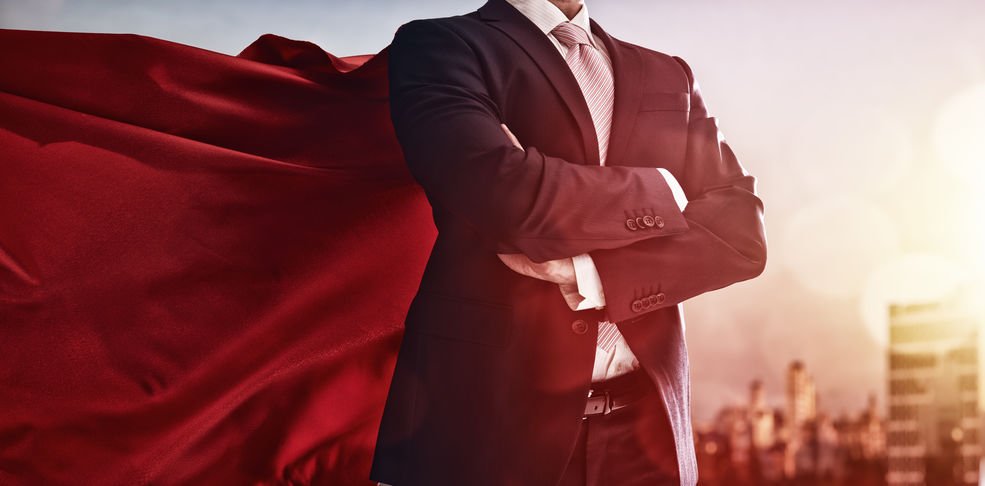 Homem vestindo roupa social, com os braços cruzados em frente ao seu peito. Ele usa também, uma capa vermelha de super-herói.