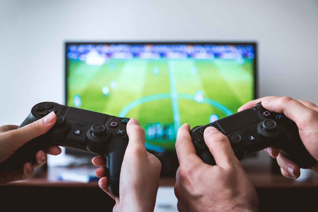 Mãos de duas pessoas segurando controles de videogame, em frente a uma televisão.