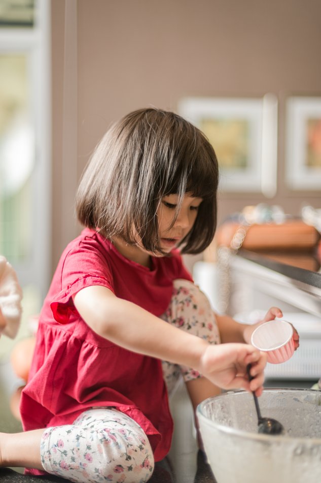 Criança em cima do balcão de uma cozinha, colocando massa de bolo em um pequeno recipiente.
