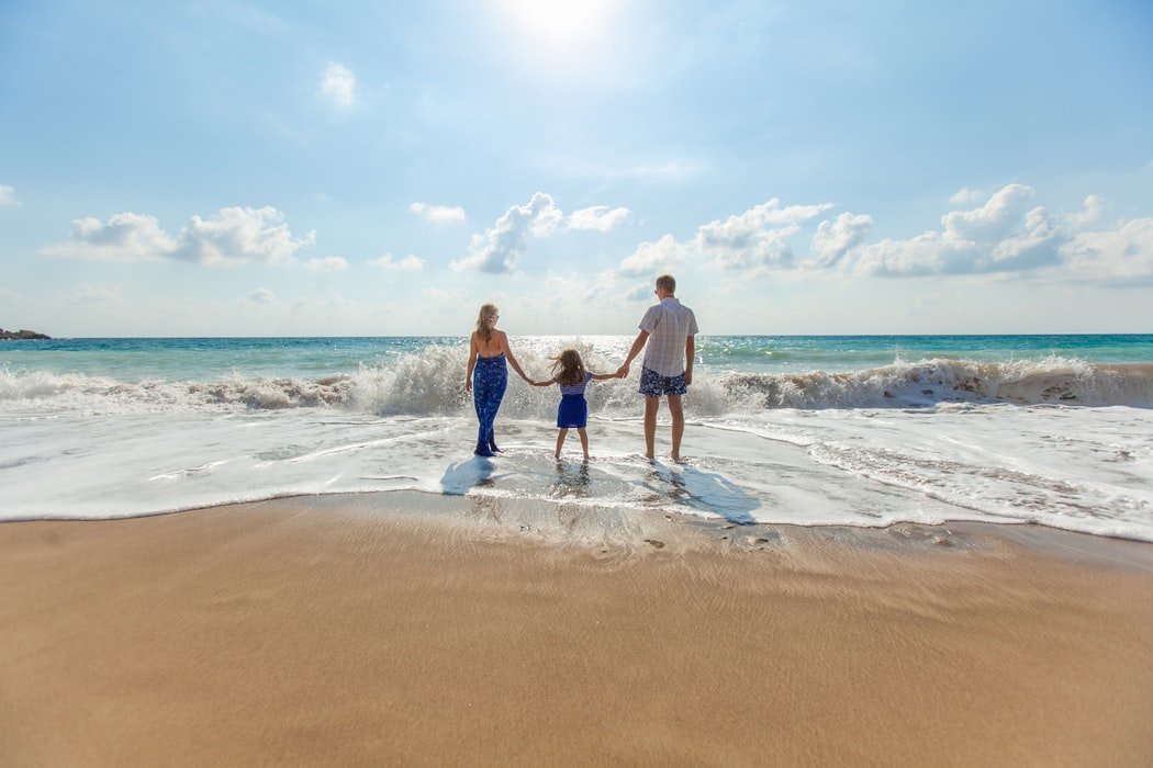 Homem, mulher e filha criança caminhando na praia em direção ao mar.