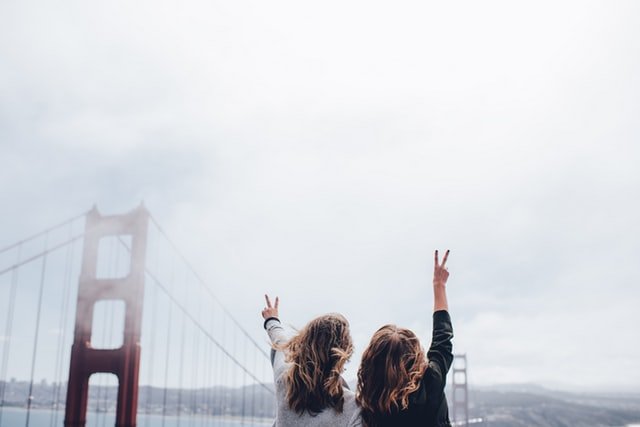 Duas mulheres de costas para a câmera, apontando seus dedos médios e indicadores para o ar, em frente a uma ponte famosa por ser ponto turístico.