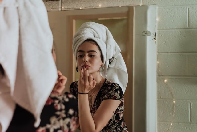 Mulher com toalha no cabelo, aplicando batom nos lábios com as mãos, de frente para um espelho.