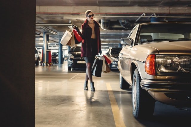 Mulher carregando muitas sacolas de compras, nas duas mãos, enquanto se aproxima de um carro em um estacionamento coberto.