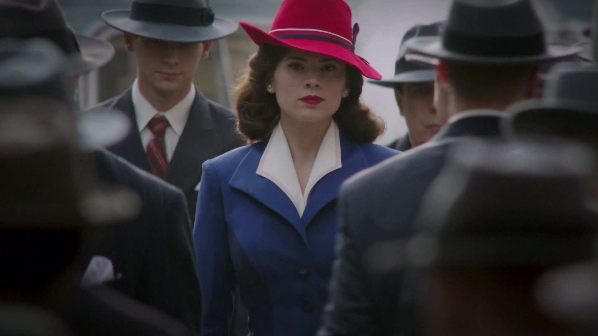 Protagonista da série andando entre multidão, com casaco azul e chapéu vermelho.