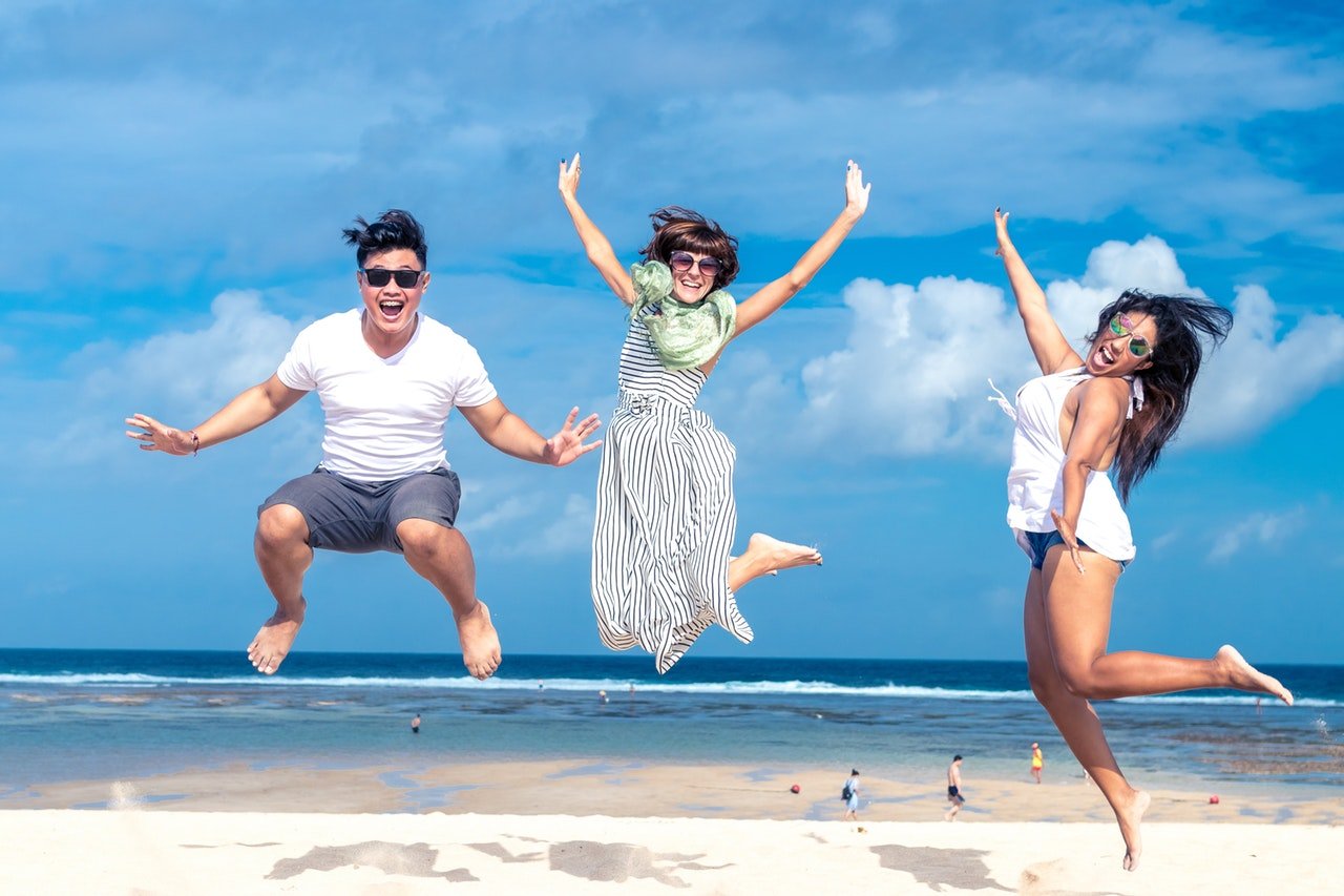 Três pessoas pulando enquanto sorriem em uma praia num dia ensolarado.