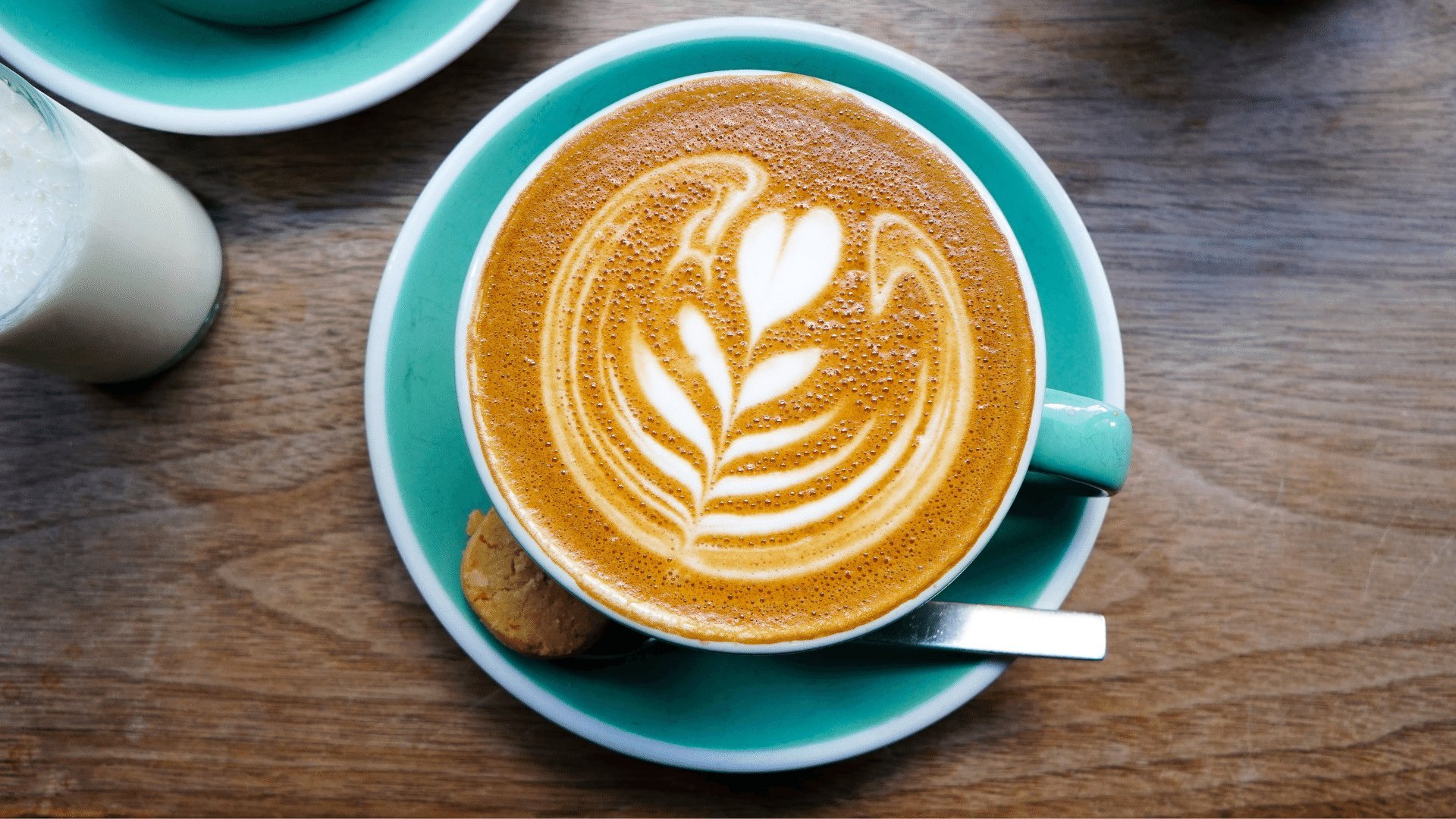 Café com um desenho de leite feito na bebida em uma xícara apoiada em um prato com um biscoito que está sobre uma superfície de madeira