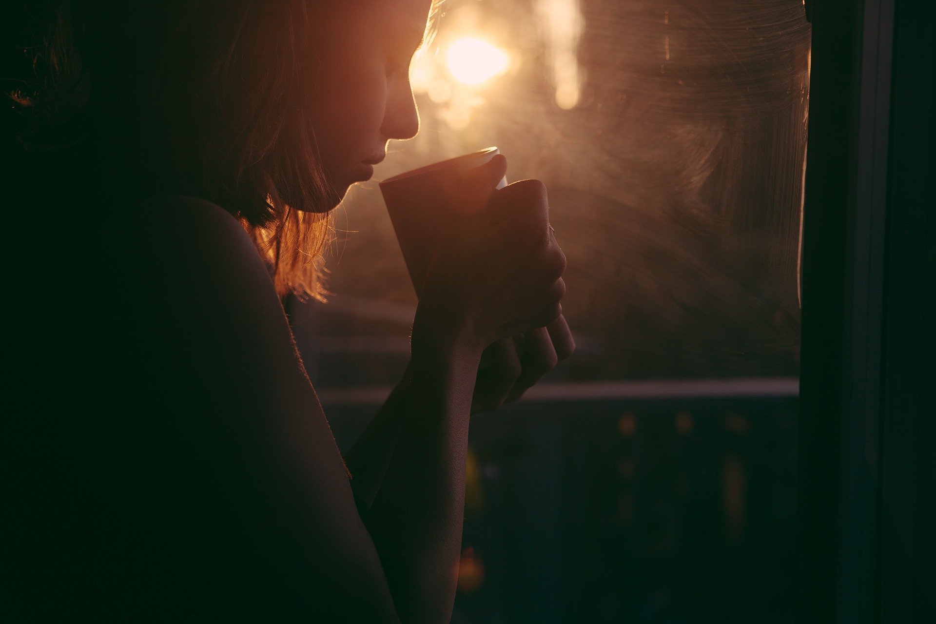 Mulher segurando xícara próximo ao rosto ao lado de uma janela