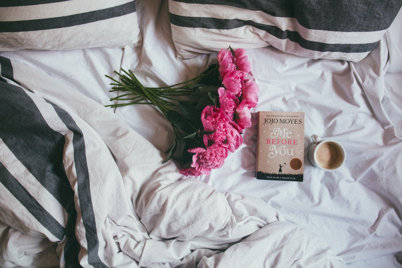 Cama com um buquê de flores rosas, um livro e uma caneca de café com leite.