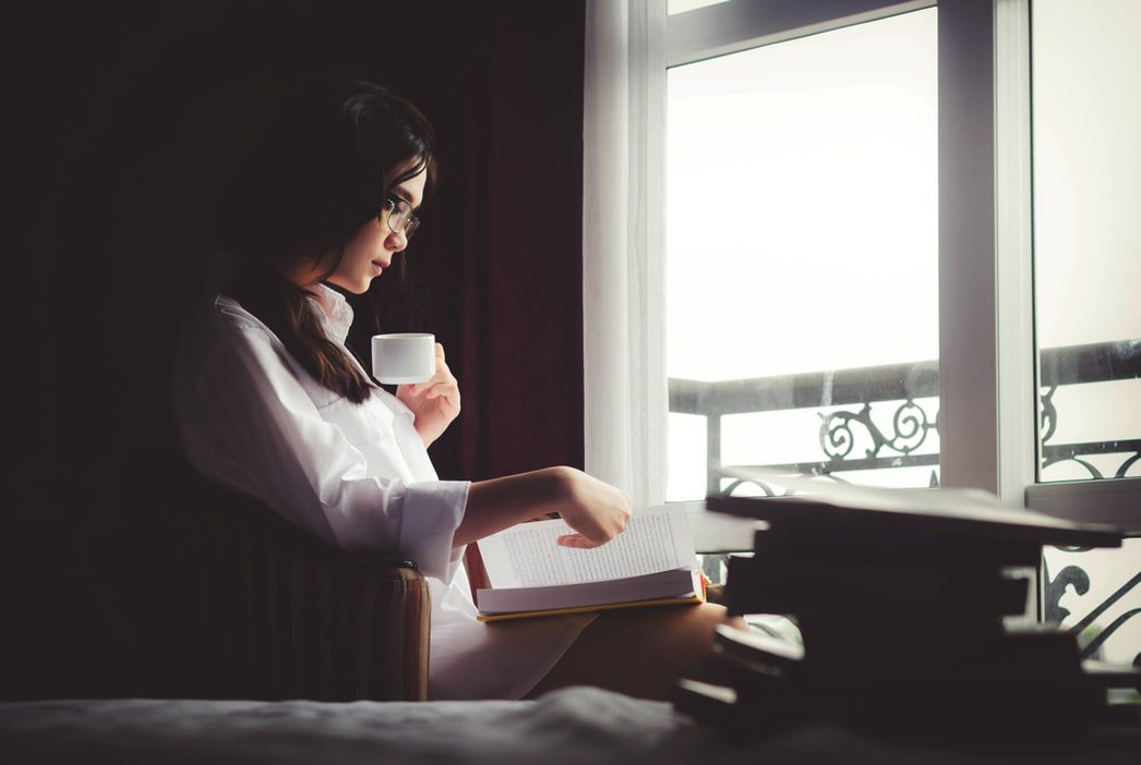 Mulher de frente para uma janela, lendo um livro e segurando uma xícara.