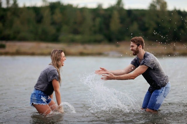 Homem e mulher dentro de um rio, com água na altura dos joelhos, vestidos com camiseta e bermuda, jogando água um no outro.