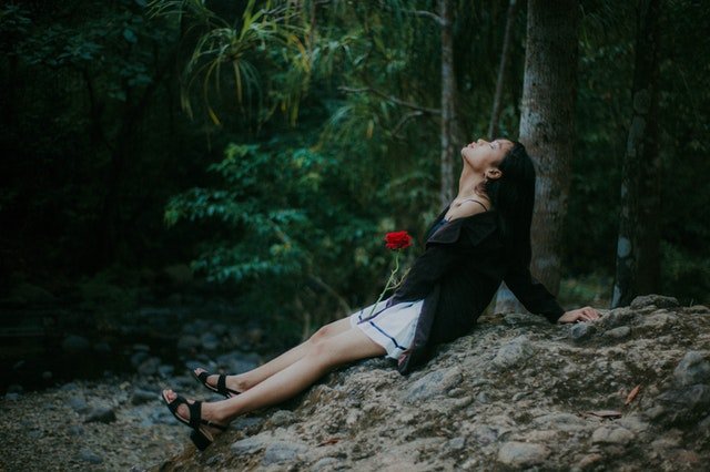Mulher sentada em pedra na natureza, segurando uma rosa vermelha no colo e com a cabeça inclinada para cima.