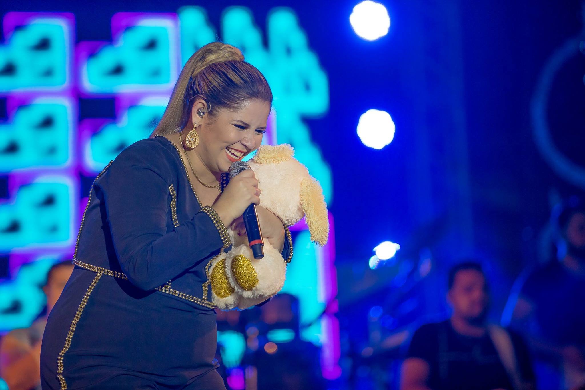 Marília Mendonça cantando feliz em show segurando um urso de pelúcia