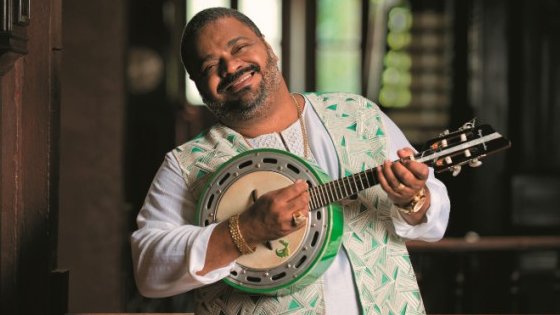 Fotografia de Arlindo Cruz sorrindo e segurando um banjo.