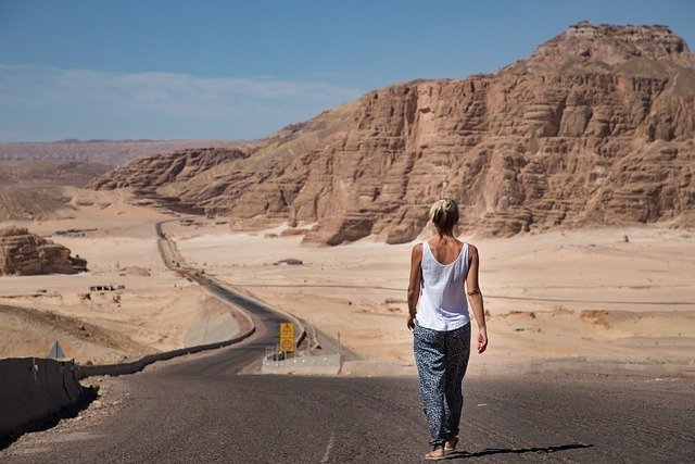 Mulher andando de chinelos em uma rodovia no meio de um deserto rochoso.
