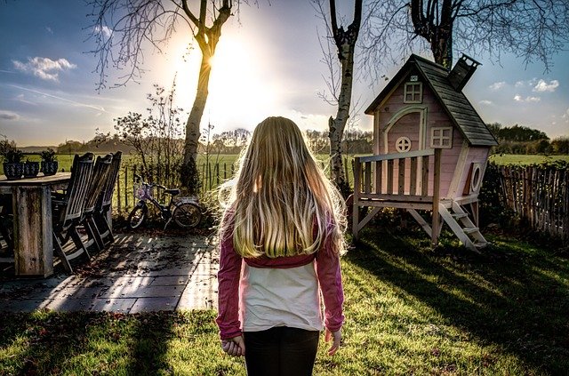 Menina de costas para a câmera, em um jardim com casa de boneca grande e mesa de madeira. Ela olha em direção ao sol.