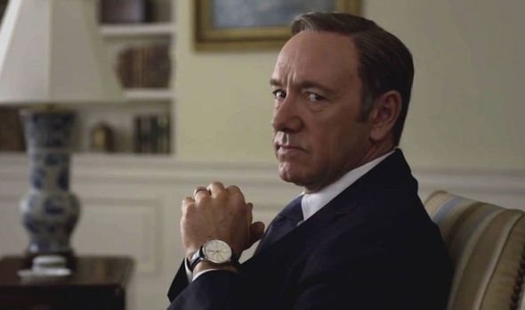 Kevin Spacey como Frank Underwood, vestindo terno e gravata, com um relógio em seu pulso esquerdo. Ele tem as mãos entrelaçadas e olha para a câmera de lado.