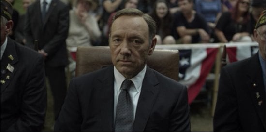 Kevin Spacey como Frank Underwood, vestindo terno e gravata, olhando desanimado para algo atrás da câmera. Ao fundo dele, diversas pessoas estão sentadas como se estivessem assistindo algo.