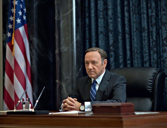 Kevin Spacey como Frank Underwood, vestindo terno e gravata, com as mãos entrelaçadas em cima de uma escrivaninha. Uma bandeira dos Estados Unidos está como decoração no fundo, ao lado esquerdo.