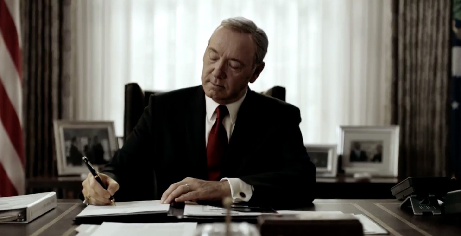 Kevin Spacey como Frank Underwood, vestindo terno e gravata, assinando um documento em seu escritório.
