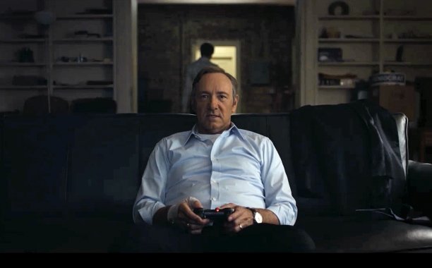 Kevin Spacey como Frank Underwood, vestindo uma camisa azul, sentado em um sofá, segurando um controle de videogame com as duas mãos.