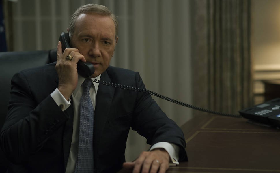 Kevin Spacey como Frank Underwood, vestindo terno e gravata, com o braço esquerdo apoiado em uma mesa, e a mão direita segurando um telefone em seu ouvido.
