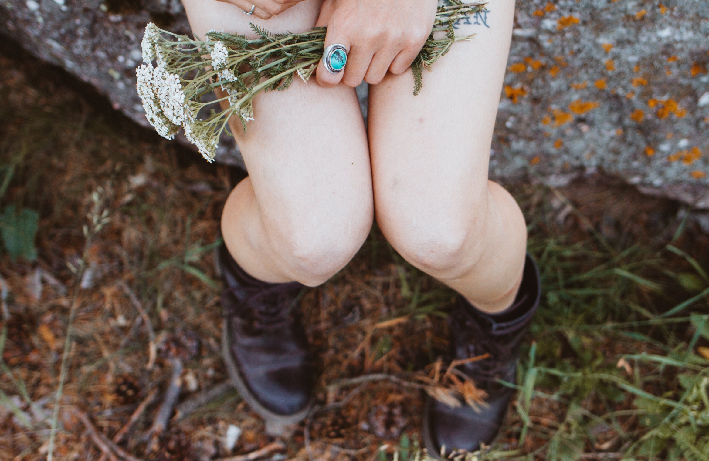 Mulher com as pernas expostas, usando coturno preto. Seus joelhos estão se tocando, e ela segura um pequeno buquê de flores em cima das coxas.
