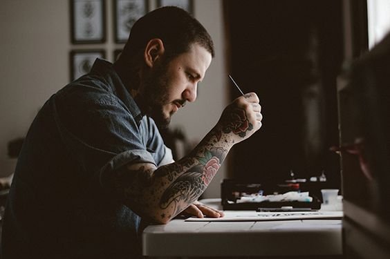 Homem sentado em frente a mesa, escrevendo algo em um papel, segurando uma caneta.