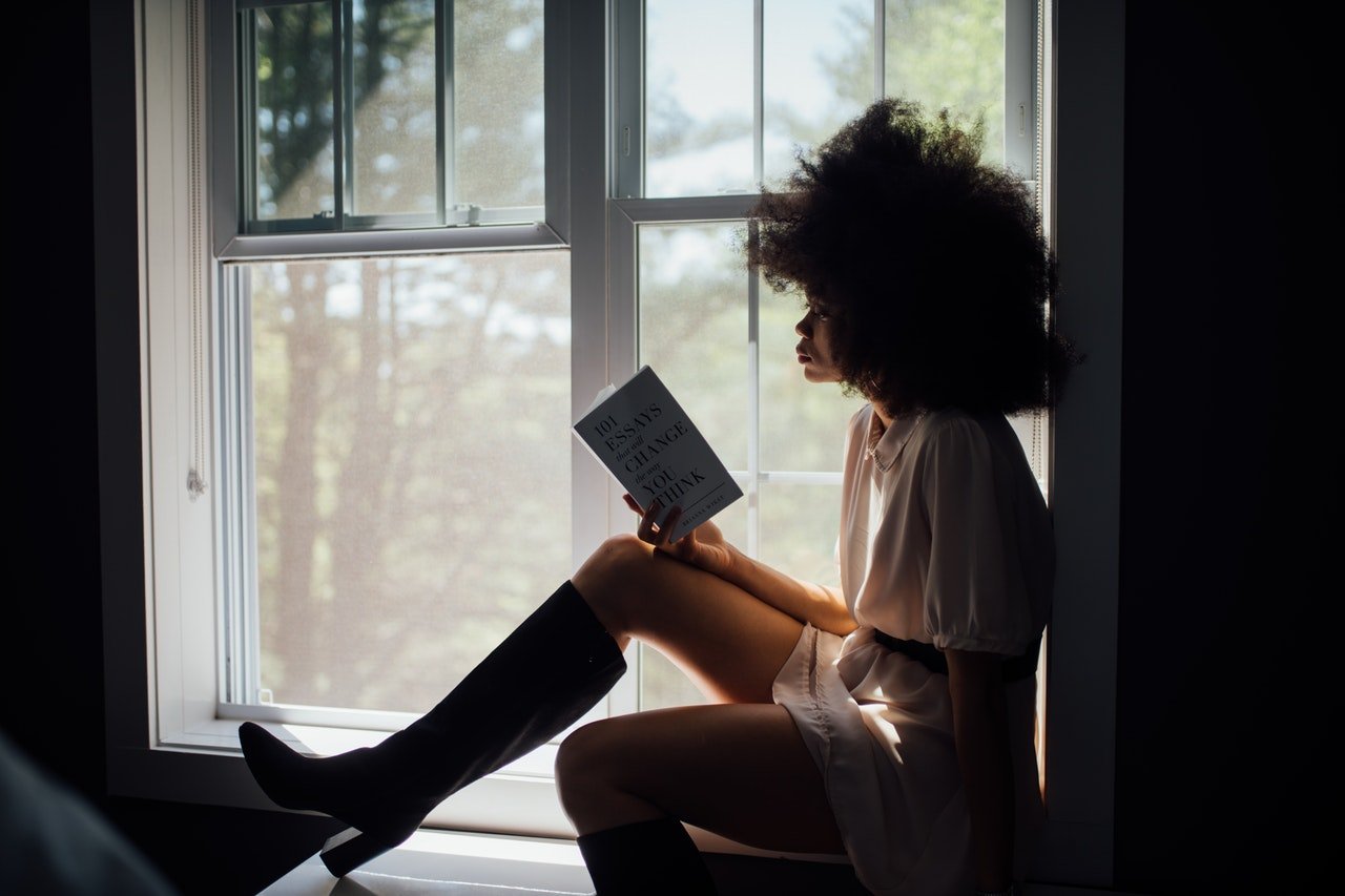 Mulher sentada em janela com uma perna esticada e a outra flexionada para baixo, lendo um livro.