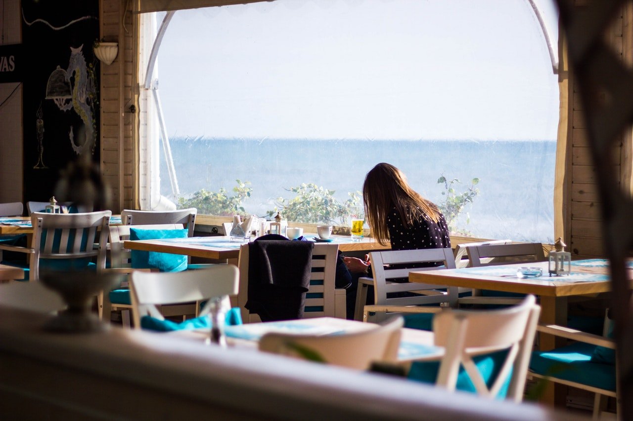 Garota sentada em um restaurante de frente para o mar, sozinha em uma mesa, e de costas para a câmera.