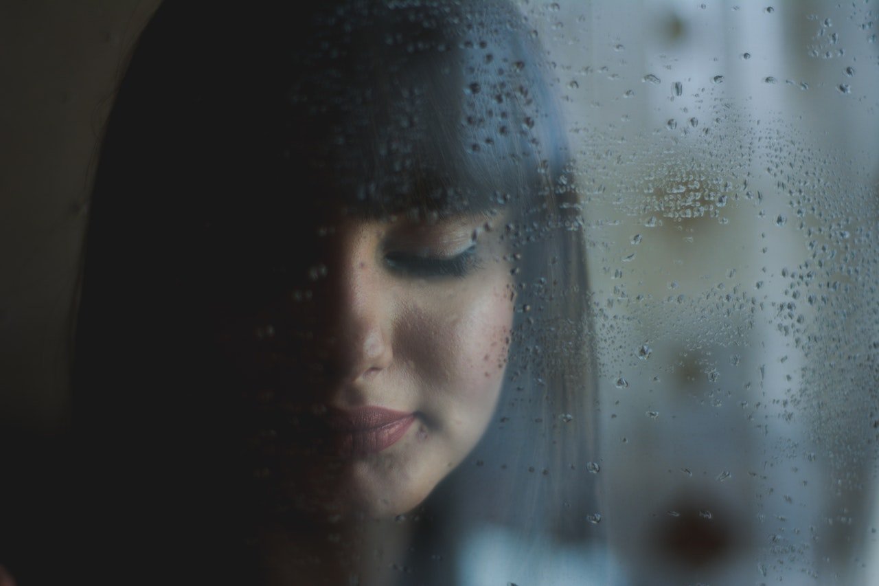 Garota com olhos fechados em frente de janela molhada de chuva.
