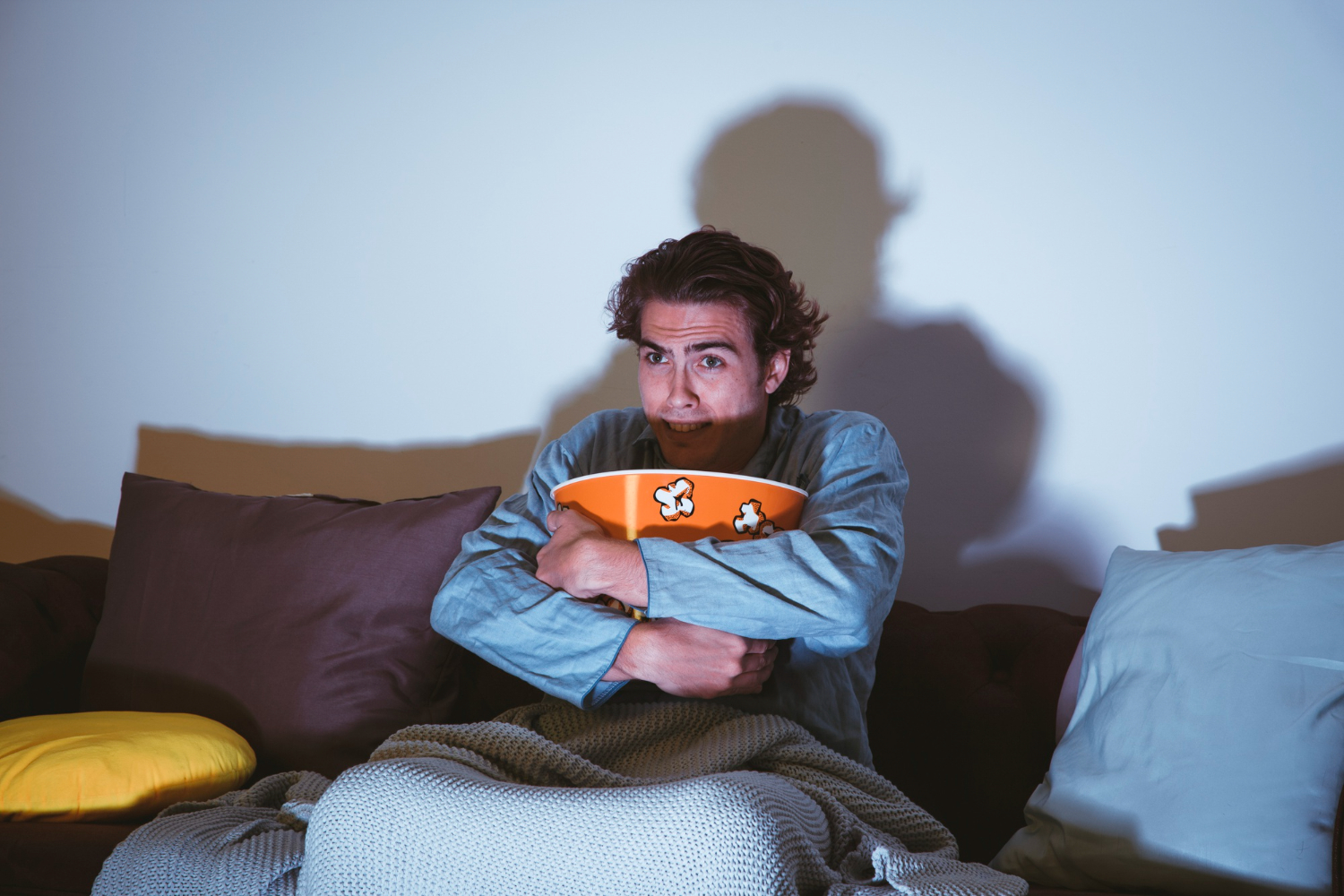 Homem sentado no sofá abraçando o balde de pipoca com expressão de medo