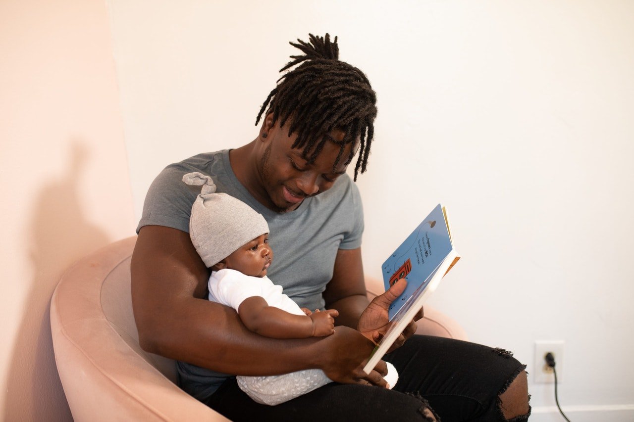 Homem olhando para bebê em seu colo, enquanto segura um livro colorido na frente dos dois.