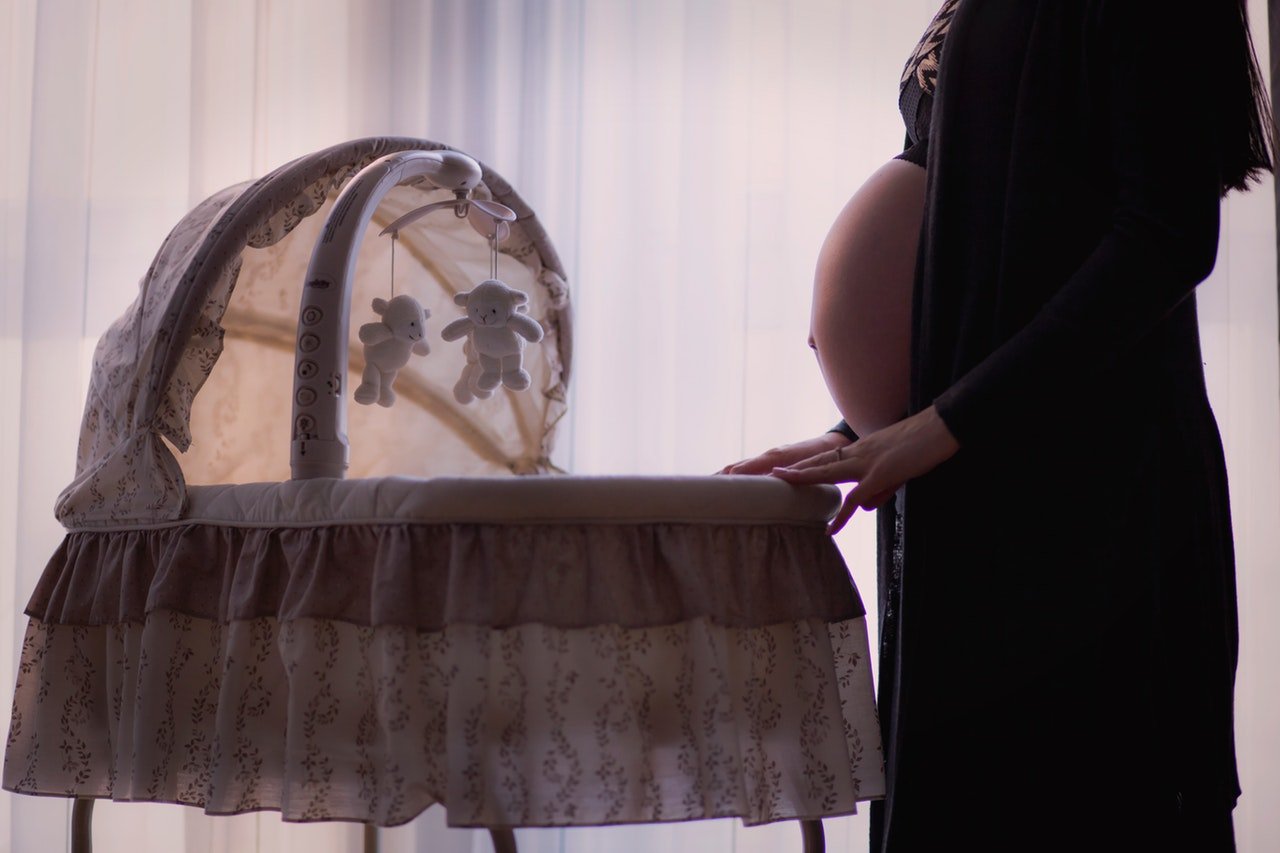 Mulher grávida usando um roupão aberto, com a barriga para fora, em frente a um pequeno berço com móbile de ursinhos.