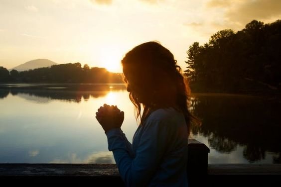 Silhueta de uma mulher orando, de perfil, à frente de um rio sob o pôr do sol.