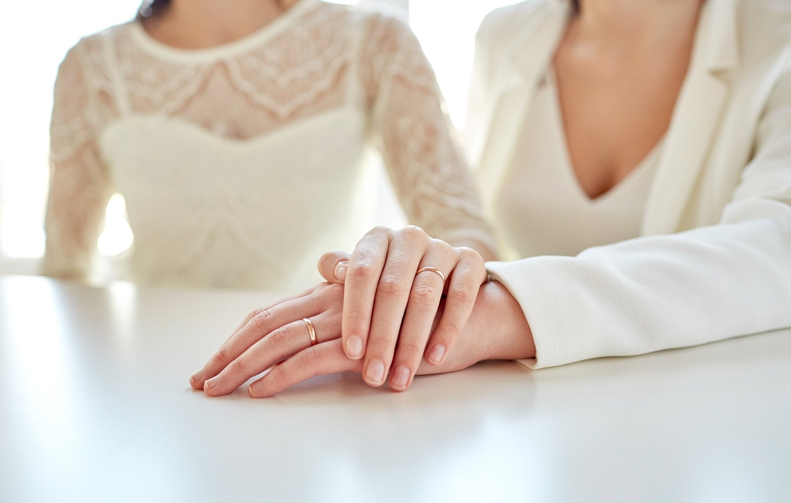 Duas mulheres com roupas brancas, com suas mãos esquerdas apoiadas em uma mesa. A mão de uma das mulheres está sobre a mão da outra.