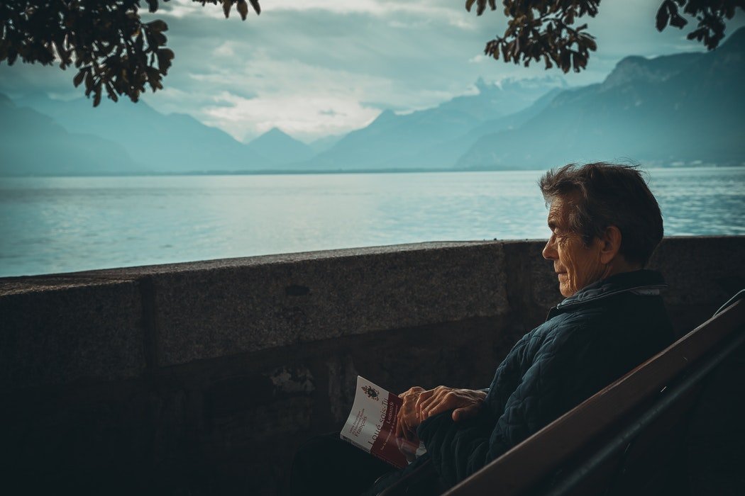 Homem idoso sentado em frente ao mar, com um livro no colo.