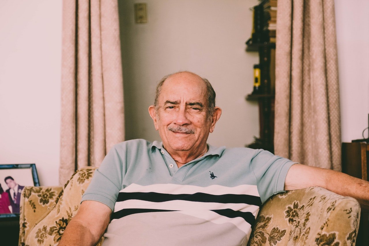 Homem idoso sentado no sofá de uma sala e sorrindo.