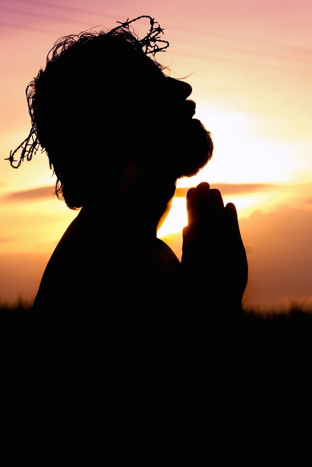 Silhueta de Jesus Cristo com mãos unidas em frente ao corpo e céu em pôr-do-sol ao fundo.