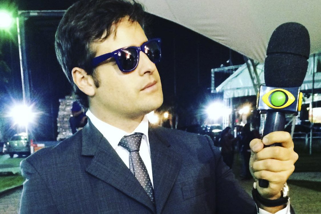 Rodrigo Scarpa, como Repórter Vesgo, no programa 'Pânico na Band', usando óculos escuro, terno, gravata e segurando um microfone.