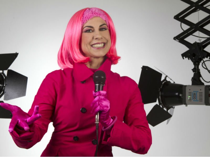Atriz Ângela Dip interpretando Penélope do 'Castelo Rá-Tim-Bum', com roupas e peruca rosa, sorrindo e segurando um microfone.