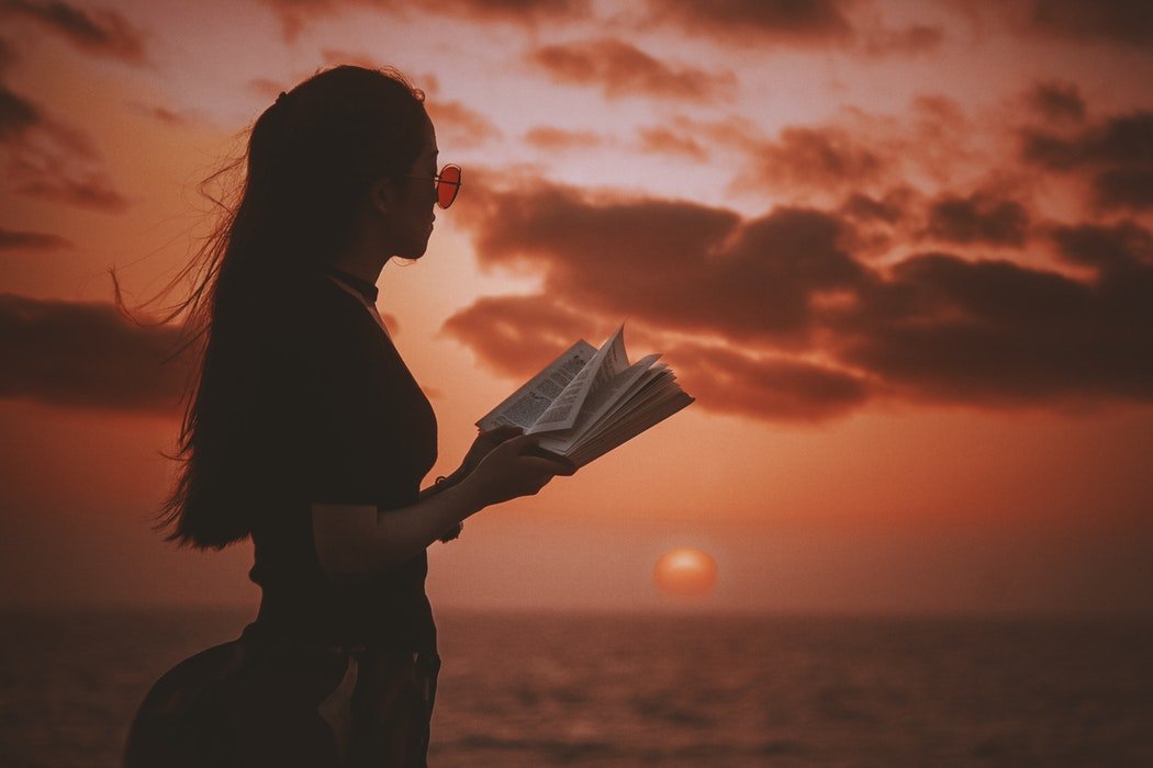 Mulher de óculos escuros lendo livro com pôr-do-sol ao fundo