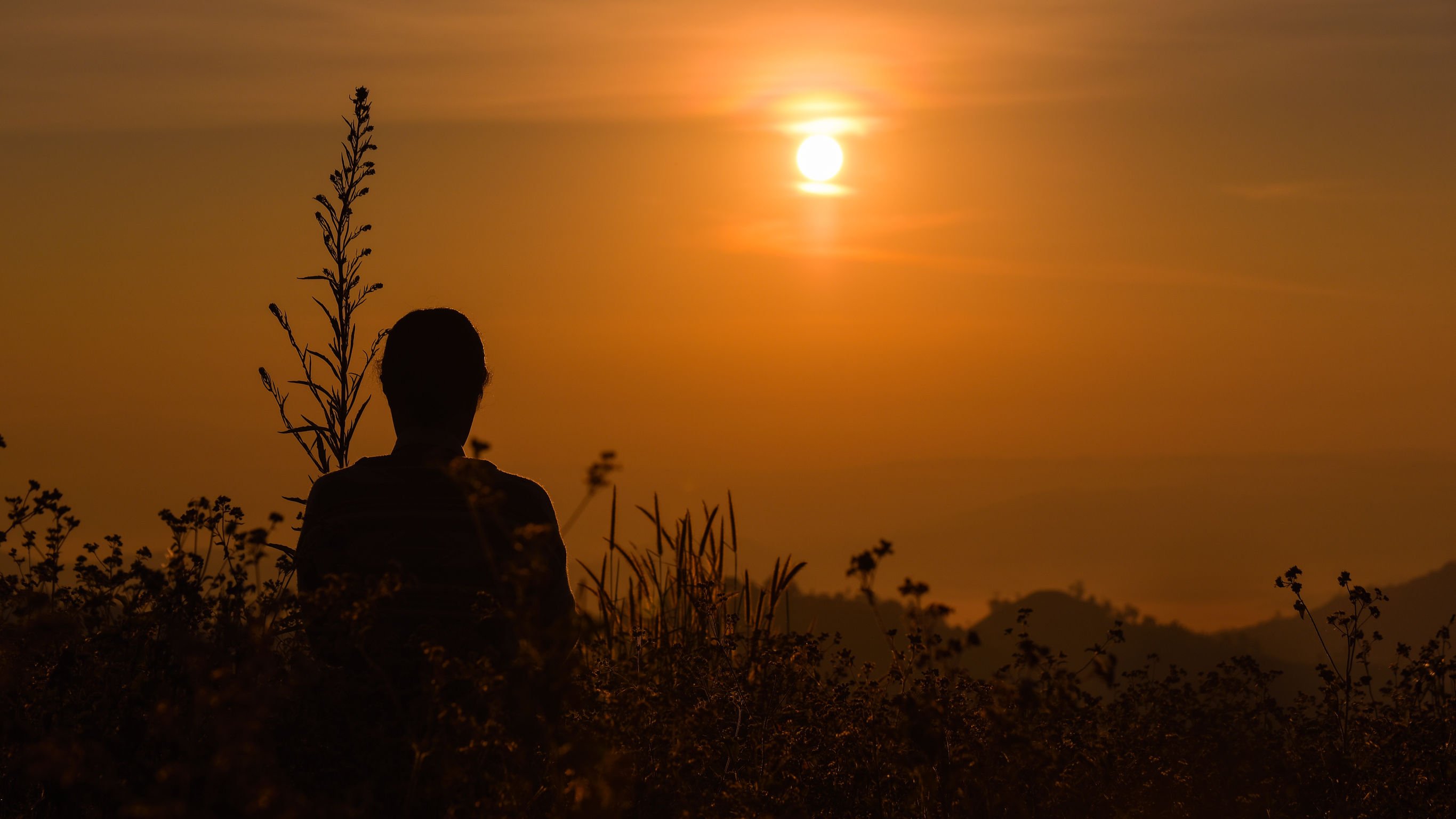 Silhueta de pessoa assistindo o pôr do sol no horizonte, sentada em uma montanha.