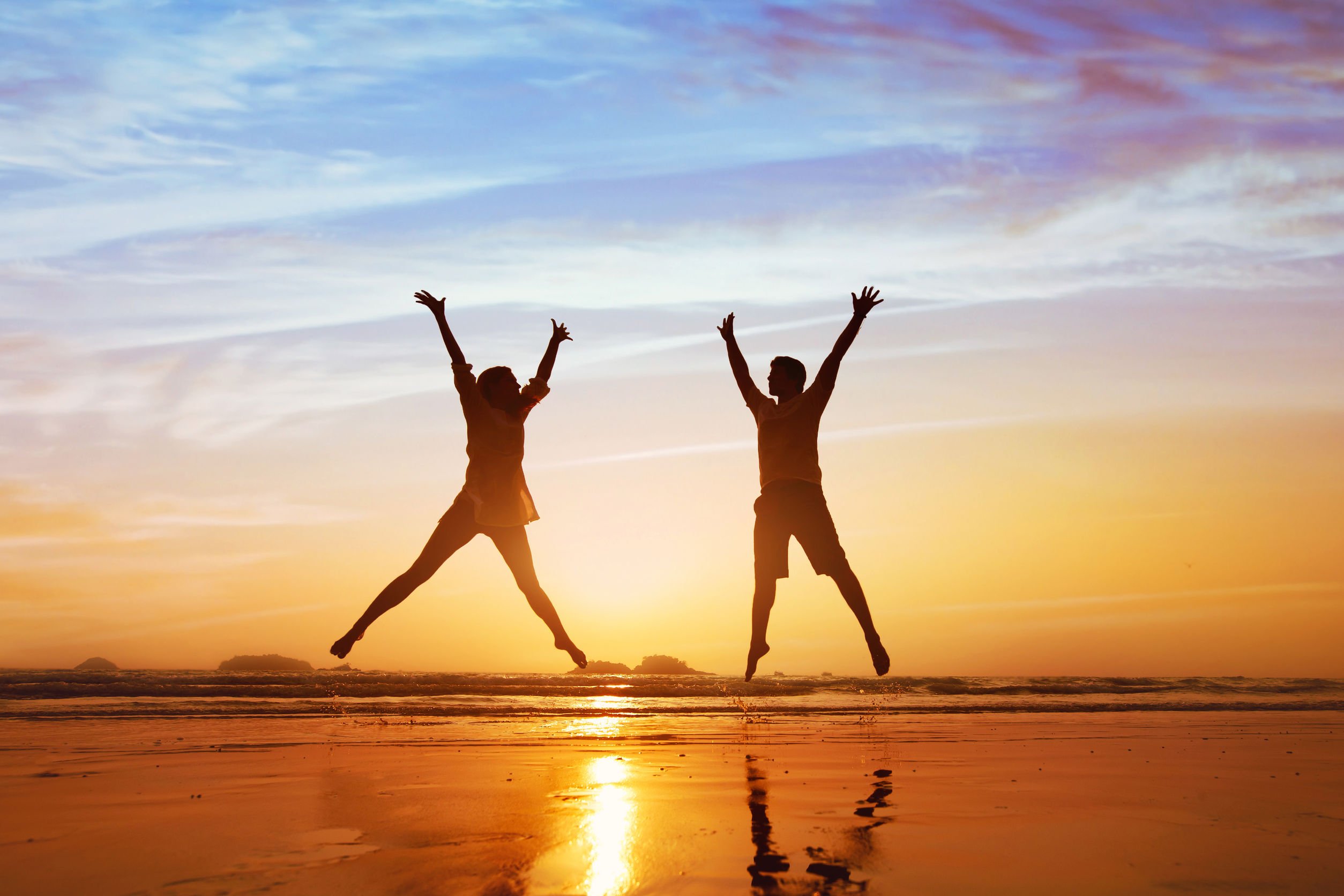 Duas pessoas pulando com os braços para o alto, em uma praia durante o pôr do sol.