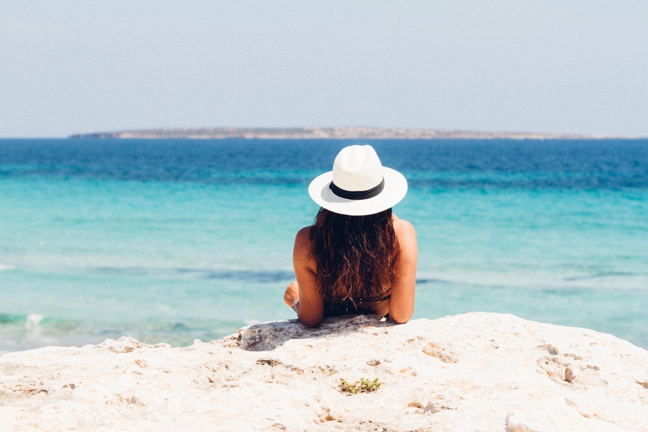 Mulher usando chapéu, deitada na areia de uma  praia, se apoiando em seus cotovelos e olhando para o mar.